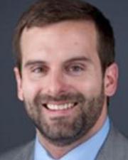 Man with dark hair and a dark beard wearing a light gray suit.