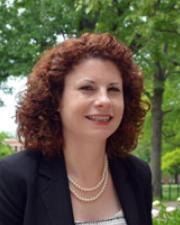 Woman with red curly hair wearing a black blazer with a pearl necklace.