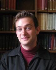 Man with wavy brown hair, wearing a red turtleneck sweater and a black jacket, standing in front of a bookshelf