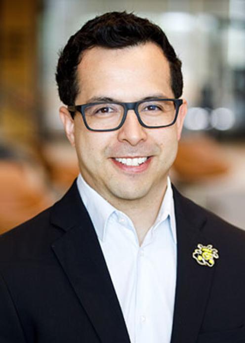 Man with short dark hair, wearing glasses, a white button-down shirt, and a black blazer with a Wichita State Shocker on the lapel