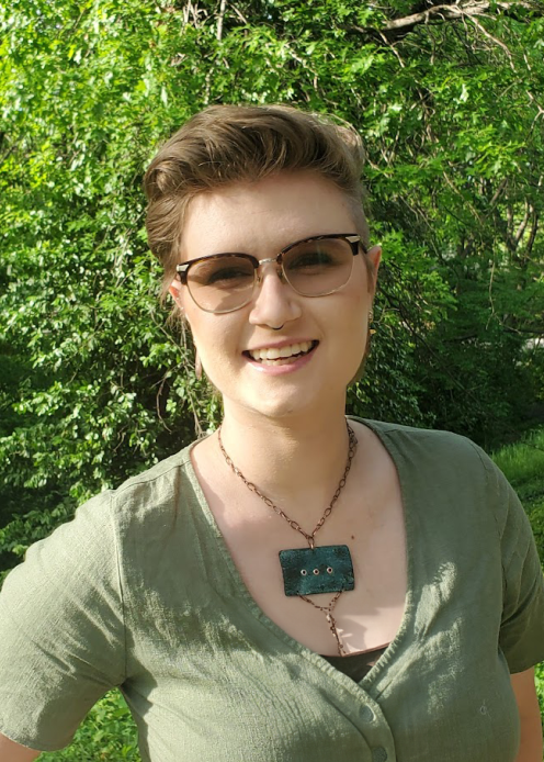 Person with light hair in a mullet wearing a green shirt and blue necklace